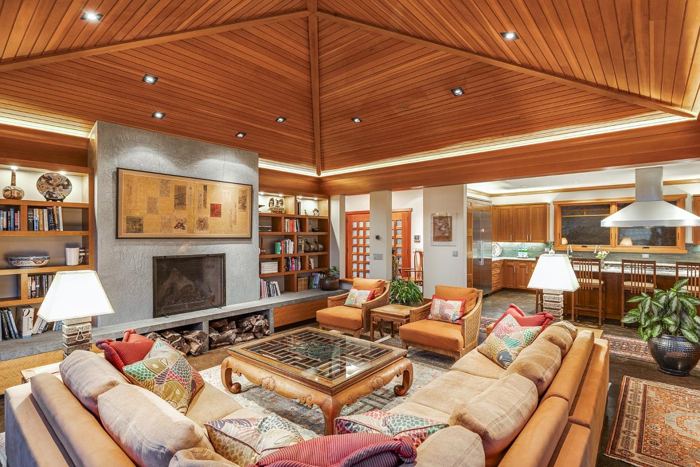 This spacious, open-concept living room features a dramatic vaulted wood-paneled ceiling and a large stone fireplace as its central focal point. The room is furnished with comfortable, warm-toned seating, a decorative glass-topped coffee table, and built-in shelving, seamlessly transitioning into an adjacent kitchen area. The overall aesthetic is a sophisticated blend of rustic warmth and refined, worldly decor, creating an inviting and luxurious atmosphere.