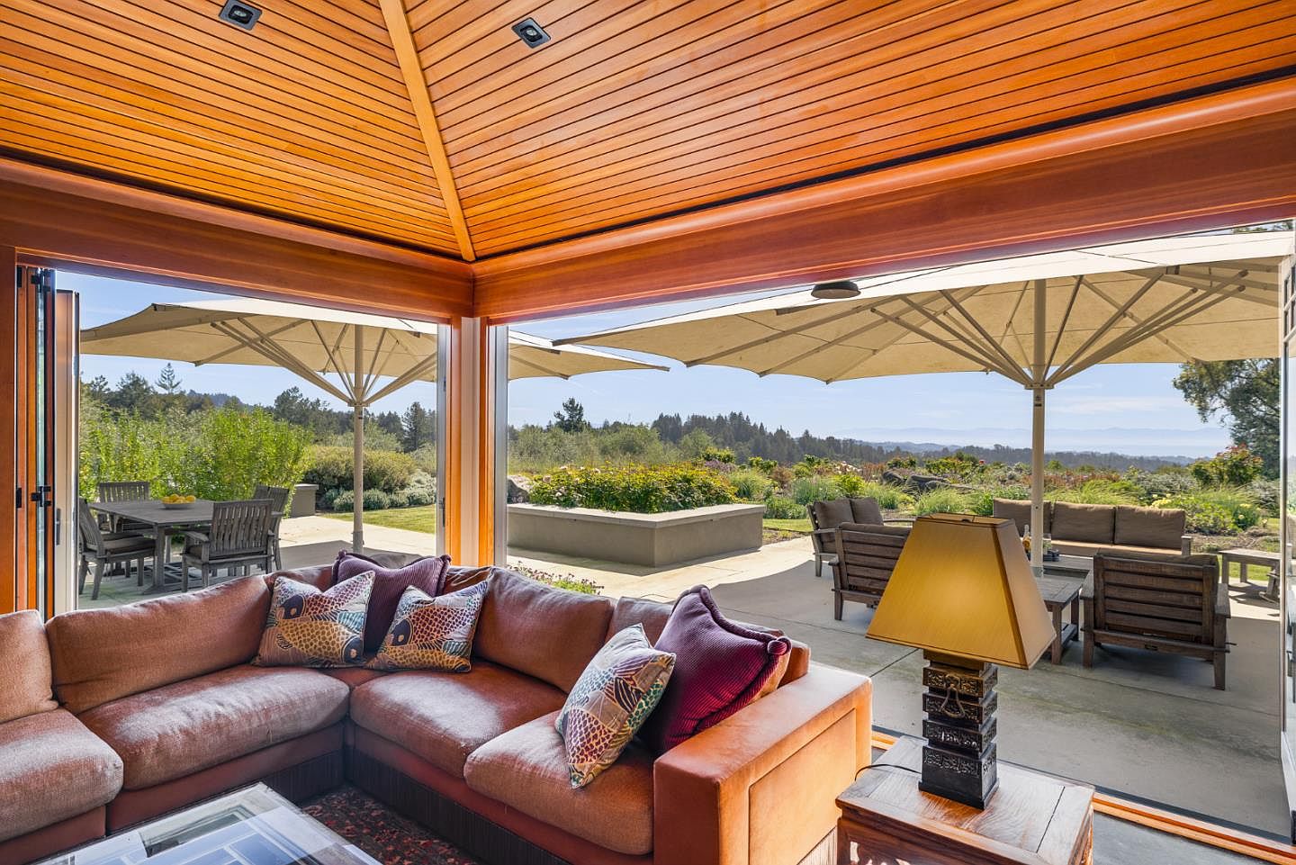 This inviting living room features a plush, L-shaped leather sofa adorned with patterned throw pillows, seamlessly transitioning to an expansive outdoor patio through large, open folding glass doors. The room is characterized by a warm, wood-paneled vaulted ceiling and a decorative table lamp, creating a cozy atmosphere that highlights the indoor-outdoor living concept. The view extends to a beautifully landscaped garden with patio furniture and large umbrellas, offering a serene and luxurious connection to nature.
