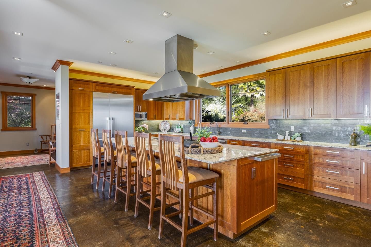 This spacious, warm-toned kitchen features custom wood cabinetry, a large granite-topped island with bar seating, and high-end stainless steel appliances. The room is anchored by a prominent stainless steel range hood and complemented by a dark, textured stone backsplash and polished concrete floors. A large window above the sink provides a scenic view of the outdoors, creating an inviting and functional space for cooking and entertaining.