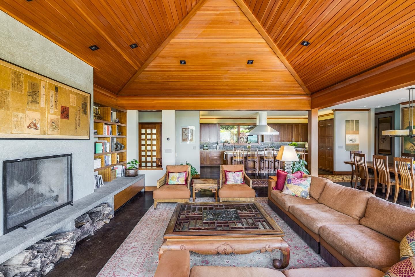 This spacious, open-concept living room features a striking vaulted wood-paneled ceiling and a large stone fireplace with built-in shelving. The warm, earthy color palette is complemented by Asian-inspired furniture, including a central coffee table and comfortable seating, creating a sophisticated and inviting atmosphere. The perspective looks from the living area toward the kitchen and dining space, highlighting the seamless flow of the home's layout.