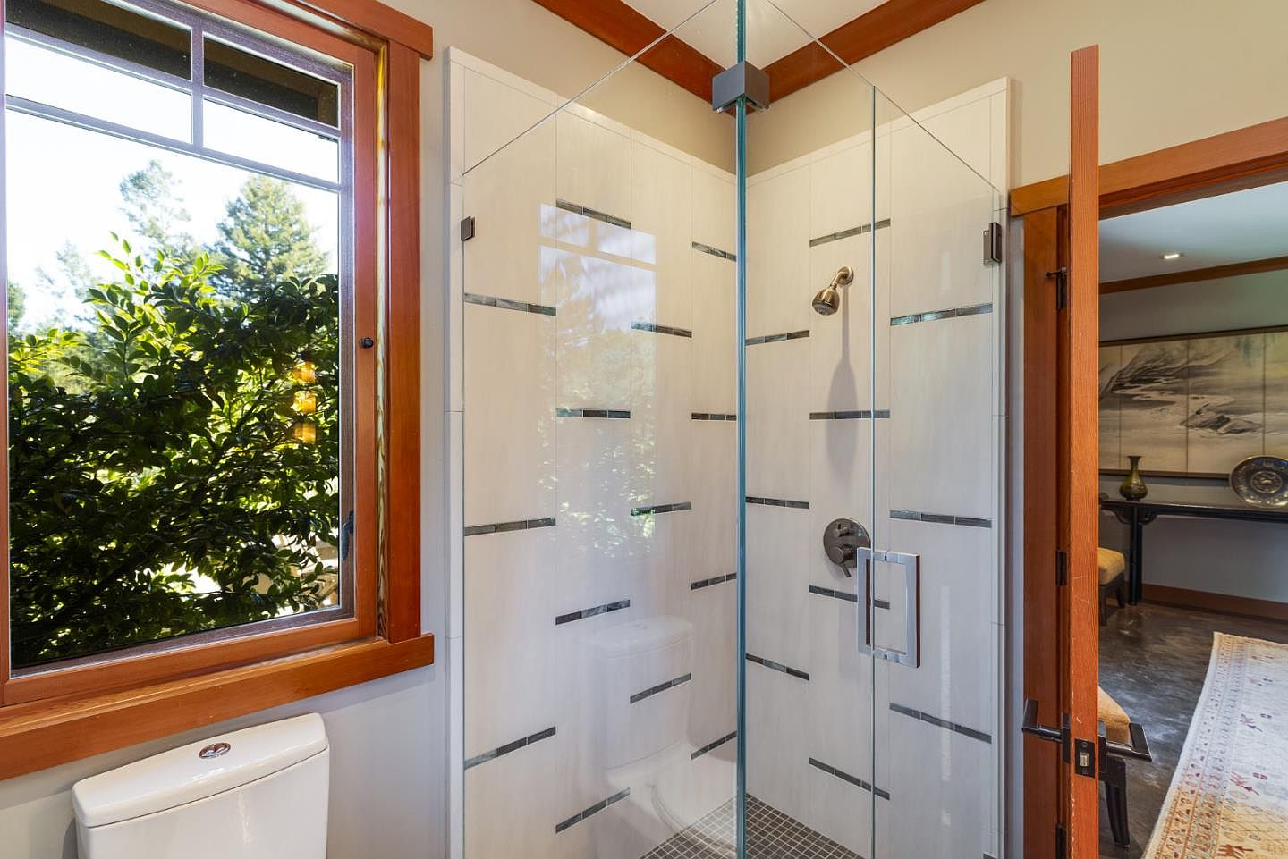 This bathroom features a sleek, glass-enclosed walk-in shower with unique horizontal tile accents and a modern showerhead. To the left, a large window with warm wood trim provides natural light and a view of lush greenery, while a doorway leads into an adjacent room with a decorative rug. The space combines contemporary design elements with natural wood finishes for a clean, inviting aesthetic.