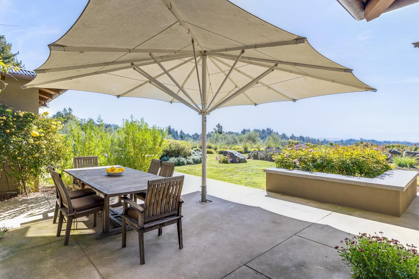 This inviting outdoor patio features a wooden dining set shaded by a large, modern cantilever umbrella, perfect for al fresco dining. The space overlooks a beautifully landscaped garden with lush greenery, vibrant flowers, and a stone planter box, creating a serene and picturesque setting. The concrete patio provides a clean, neutral foundation that emphasizes the natural beauty of the surrounding landscape.