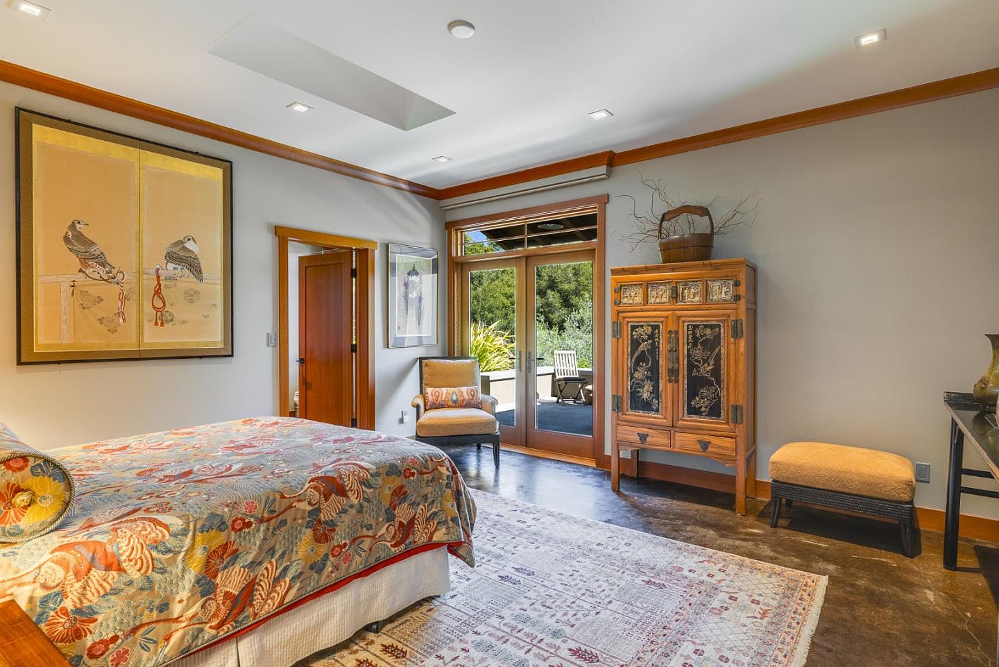 This serene primary bedroom features a sophisticated blend of Asian-inspired decor, including a large framed artwork of birds and an ornate wooden cabinet. The room is anchored by a patterned bedspread and a textured area rug, while glass doors provide a seamless transition to an outdoor patio. The space is bathed in natural light, creating a tranquil and elegant atmosphere.