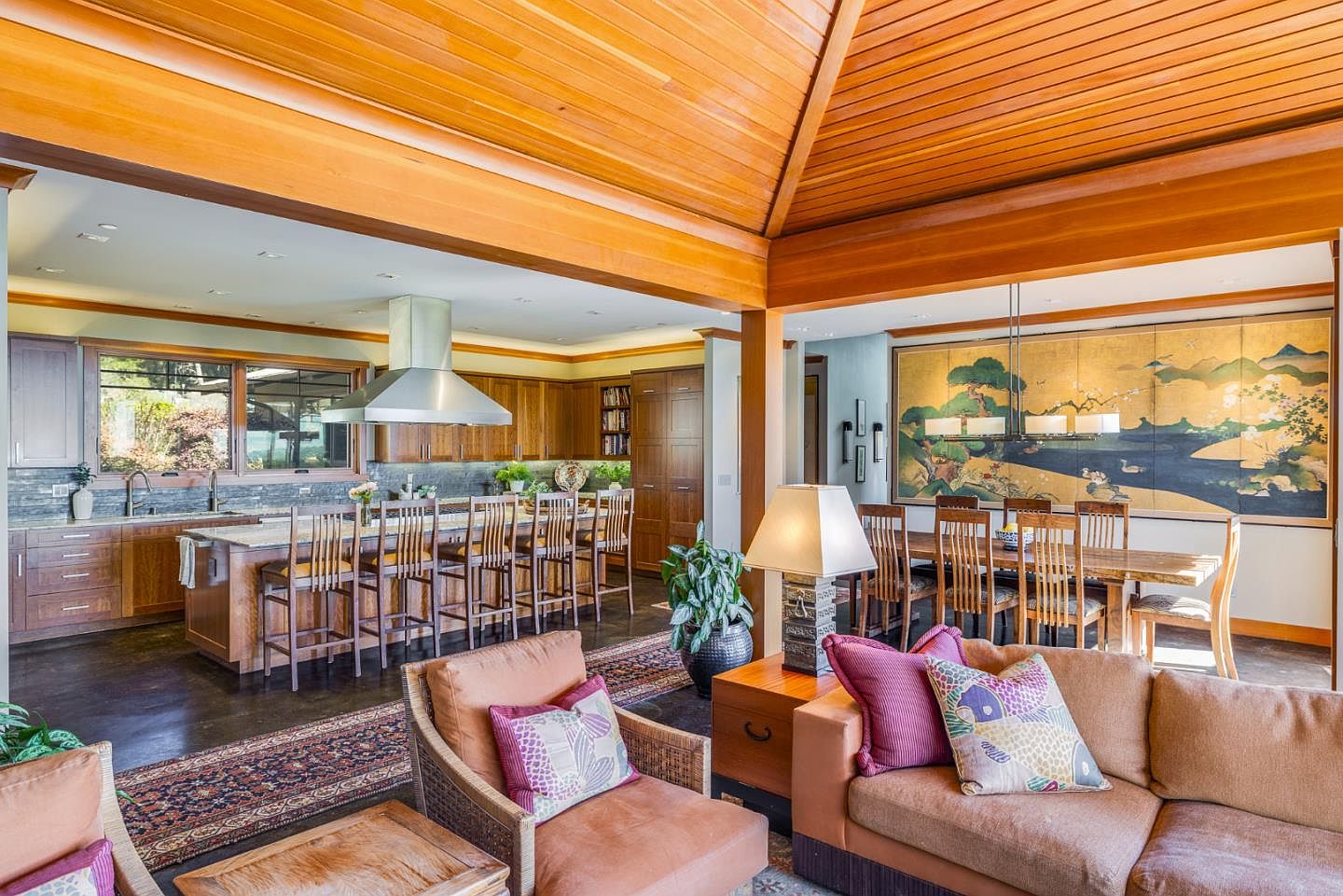 This expansive open-concept living space features a warm, wood-paneled vaulted ceiling and a seamless flow between the living, dining, and kitchen areas. The room is anchored by a comfortable seating area with plush furniture, leading into a gourmet kitchen with a large island and a formal dining space adorned with a striking landscape mural. The warm wood tones and natural light create an inviting, sophisticated atmosphere perfect for entertaining.