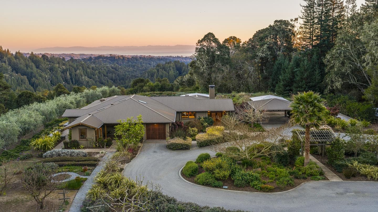 This stunning aerial view captures a sprawling, single-story residence nestled within a lush, hilly landscape at sunset. The property features a circular driveway, a detached carport, and meticulously landscaped gardens, all set against a breathtaking backdrop of rolling mountains and a distant coastline. The home's warm, earthy tones and metal roof blend harmoniously with the natural surroundings, creating an inviting and serene atmosphere.
