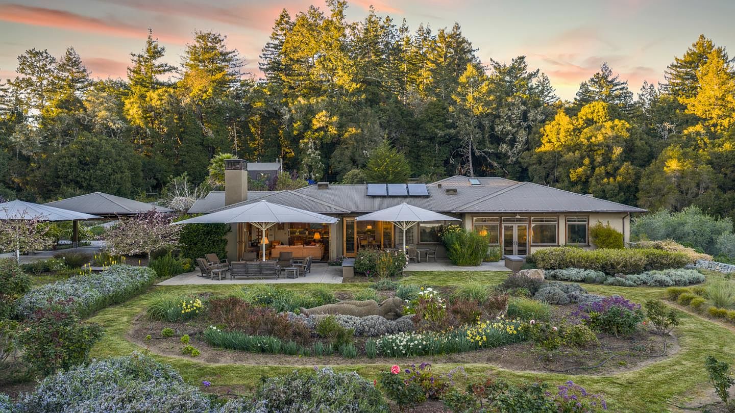 This stunning rear view of a single-story residence showcases a seamless blend of indoor-outdoor living, featuring a spacious patio shaded by large umbrellas and a beautifully landscaped garden. The home is nestled against a backdrop of lush, mature trees under a soft, golden-hour sky, highlighting its tranquil and private setting. The architectural style is understated and elegant, emphasizing comfort and connection to the natural surroundings.