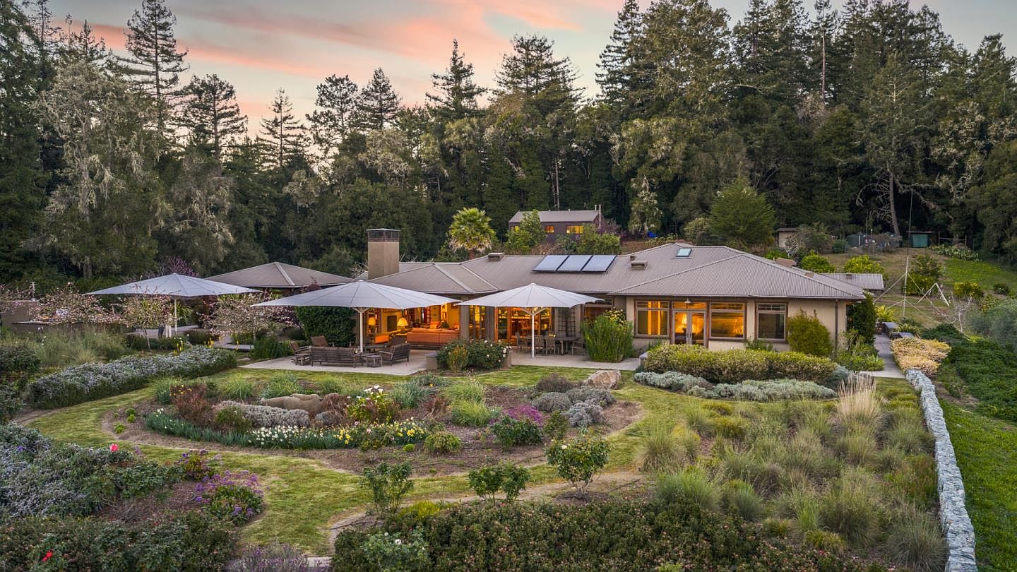 This stunning aerial perspective captures the rear of a sprawling, single-story residence nestled against a lush, forested hillside at dusk. The home features a spacious stone patio shaded by large cantilevered umbrellas, overlooking a meticulously landscaped garden filled with diverse plantings and a stone retaining wall. The warm glow from the interior windows creates an inviting atmosphere, highlighting the seamless transition between the indoor living spaces and the serene outdoor environment.