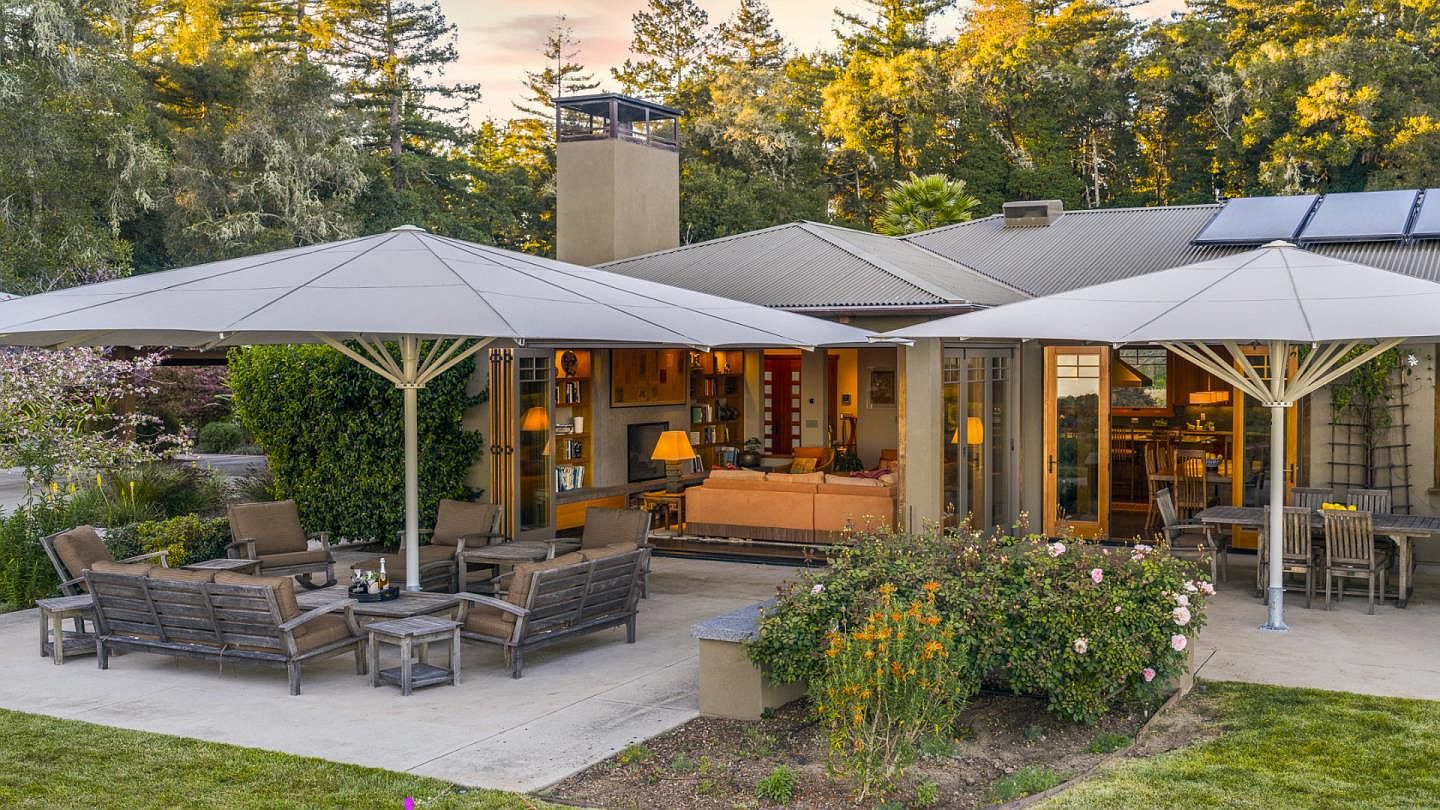 This inviting outdoor living space features a spacious concrete patio shaded by two large, modern cantilever umbrellas, perfect for al fresco dining and relaxation. The area is furnished with comfortable wooden lounge seating and a dining set, seamlessly connecting to the home's interior living room through wide glass doors. Surrounded by lush greenery and mature trees, the setting offers a serene and private atmosphere ideal for entertaining.