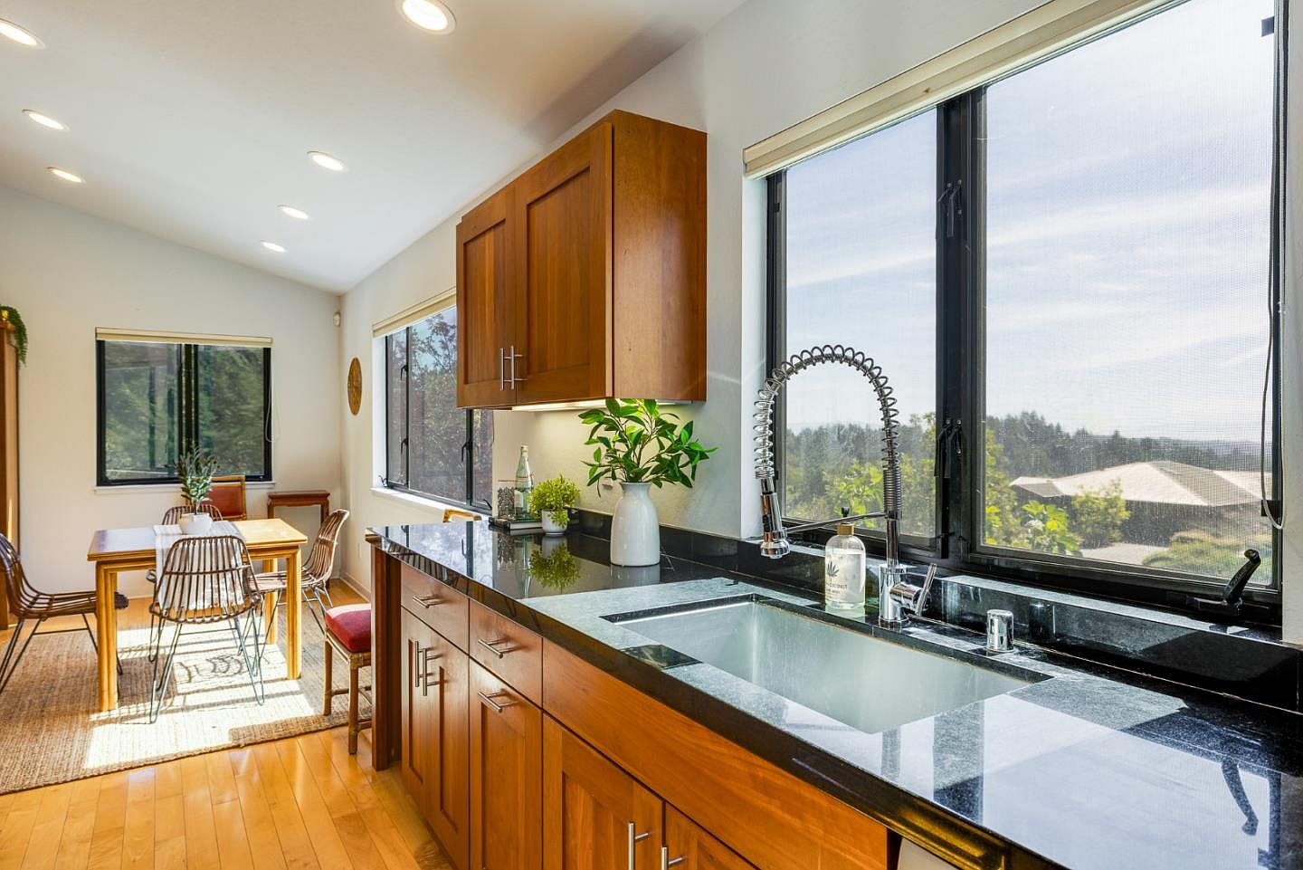 This bright and inviting kitchen features warm wood cabinetry paired with sleek black granite countertops and a modern stainless steel farmhouse sink. A large window above the sink provides a scenic view of the outdoors, while an adjacent dining area with a wooden table and stylish chairs creates a cohesive, open-concept living space. The room is illuminated by recessed lighting and natural light, offering a clean, contemporary aesthetic perfect for family living.