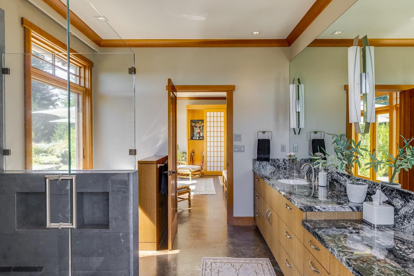 This primary bathroom features a sophisticated blend of natural wood cabinetry and striking black-and-white granite countertops. A glass-enclosed shower with dark stone tile sits to the left, while a doorway leads into a serene, light-filled adjacent room with shoji-style screens. The space is illuminated by modern vertical wall sconces and natural light streaming through the window, creating a clean, spa-like atmosphere.