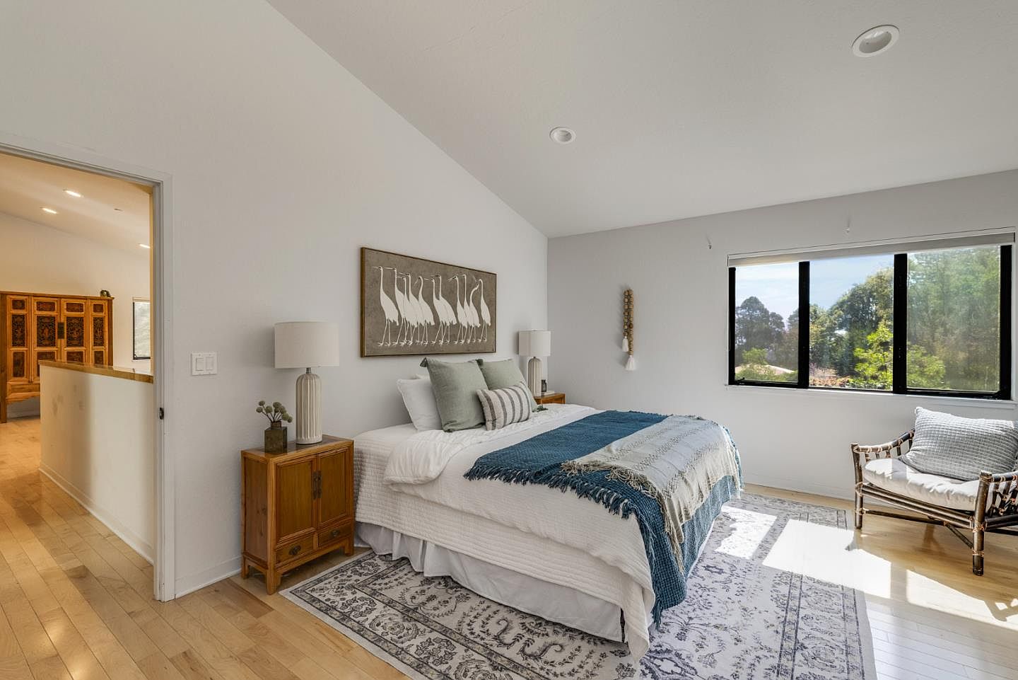 This bright and airy primary bedroom features a vaulted ceiling, a plush bed with layered blue and white bedding, and a decorative crane-themed wall art piece. A cozy rattan armchair sits by the large window, which provides ample natural light and a view of the surrounding greenery. The room is accented by a wooden nightstand and a patterned area rug, creating a serene and inviting atmosphere.