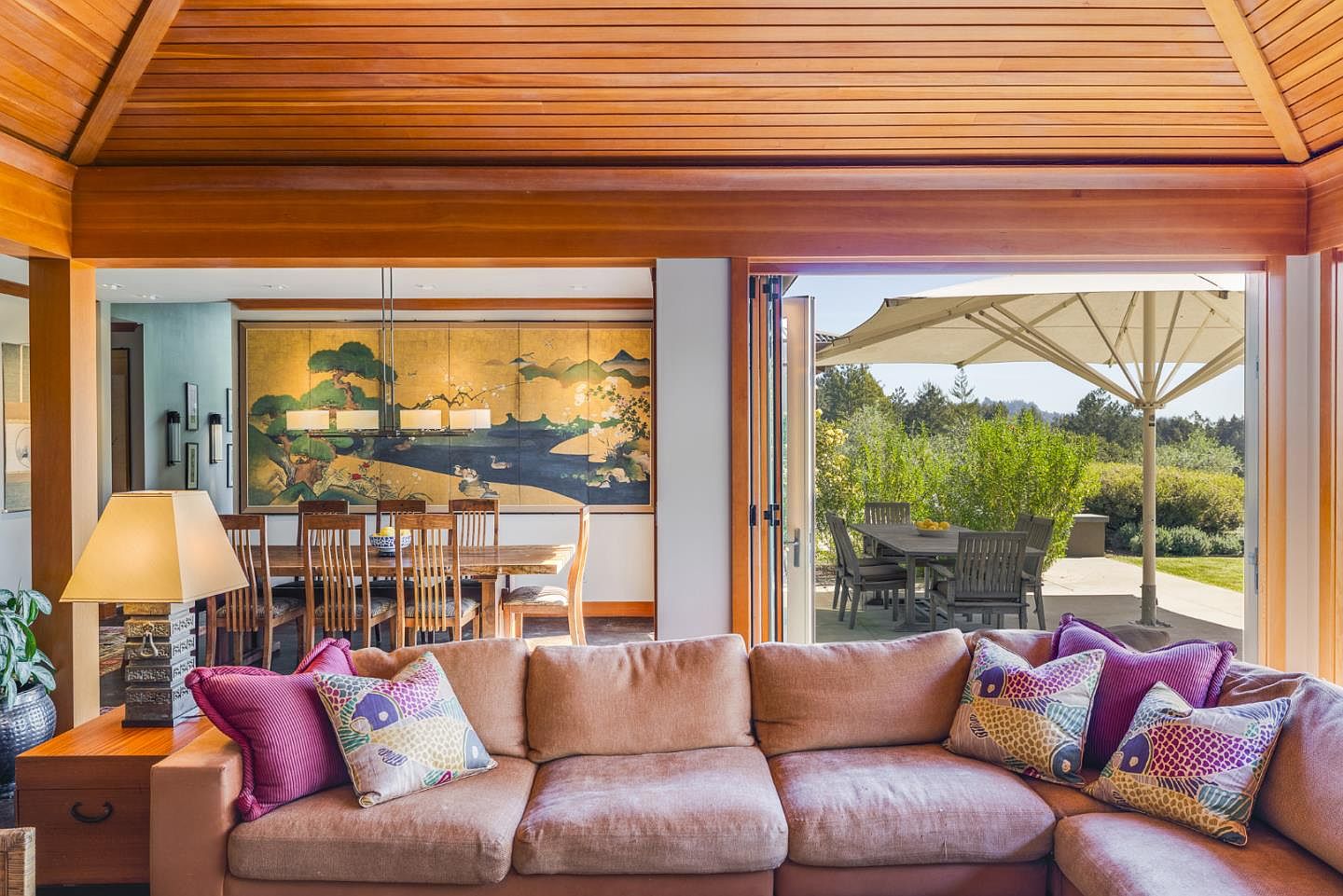 This inviting living room features a large, plush sectional sofa adorned with colorful, patterned throw pillows, set against a backdrop of warm wood-paneled ceilings and beams. The space seamlessly transitions into an elegant dining area with a traditional Japanese-style screen mural and opens through folding glass doors to a sun-drenched patio with outdoor dining. The overall aesthetic blends contemporary comfort with sophisticated, nature-inspired design elements, creating a serene and airy atmosphere.