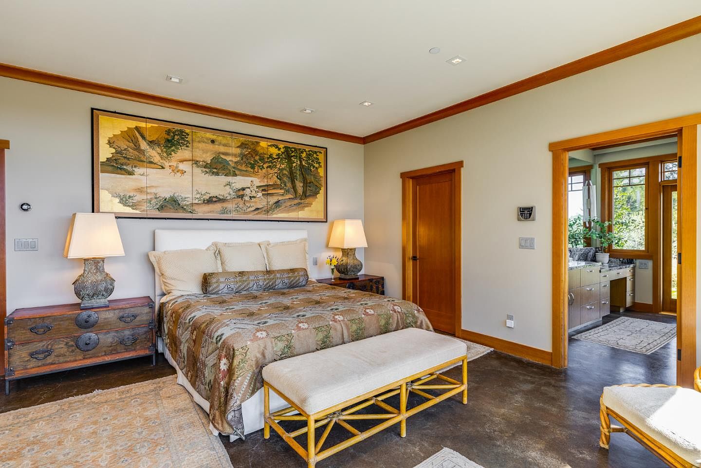 This elegant primary bedroom features a sophisticated blend of Asian-inspired decor and modern architectural elements, centered around a large, ornate landscape painting above the headboard. The room is furnished with a plush bed, a rustic wooden chest, and a woven bench, all set against a neutral color palette with warm wood trim. A doorway leads into an adjoining bathroom, creating a seamless flow and highlighting the spacious, well-lit layout of the suite.