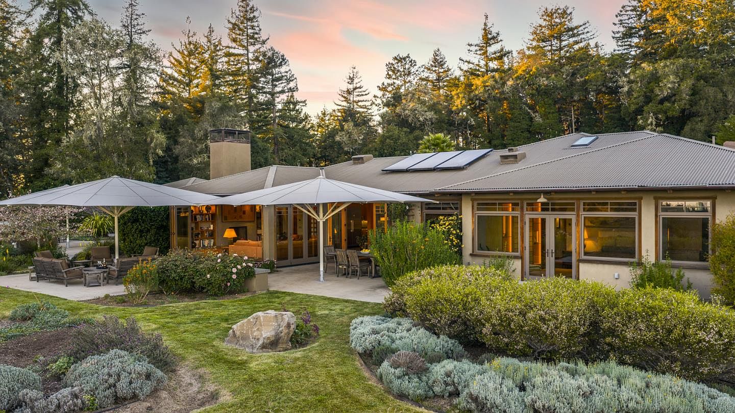 This serene rear view of a single-story residence showcases a spacious stone patio shaded by two large, modern cantilever umbrellas, perfect for outdoor entertaining. The home features a metal roof with integrated solar panels, large glass doors that blend indoor and outdoor living, and a warm, earthy exterior palette. The property is beautifully landscaped with lush green lawns, manicured shrubs, and a backdrop of towering redwood trees, creating a peaceful and private retreat.