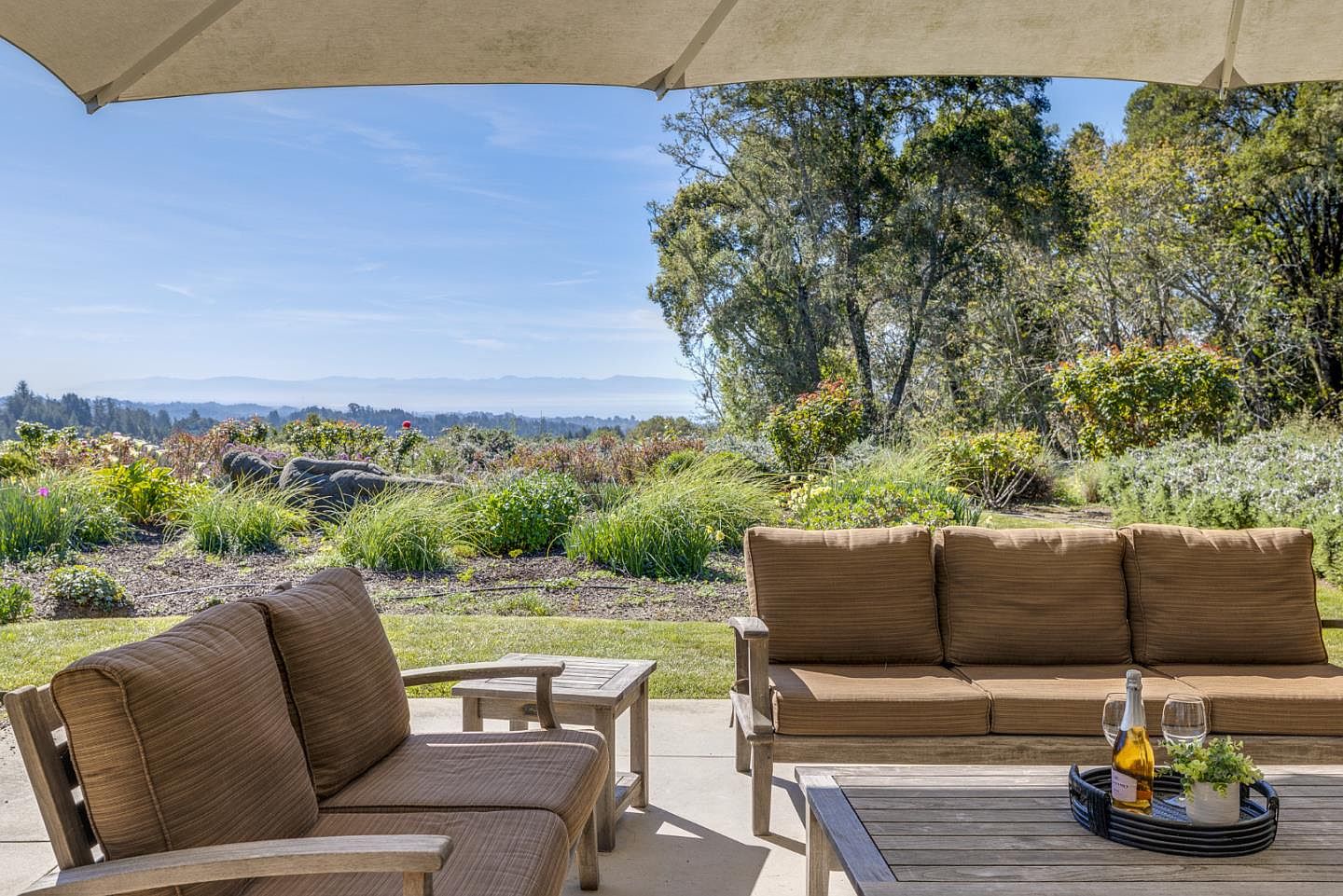 This inviting outdoor patio space features comfortable brown cushioned seating arranged around a wooden coffee table, complete with a bottle of champagne and glasses. The perspective looks out from under a shaded canopy toward a lush, expansive garden and a breathtaking, panoramic view of distant mountains under a clear blue sky. The setting offers a serene and luxurious atmosphere, perfect for relaxation and entertaining.