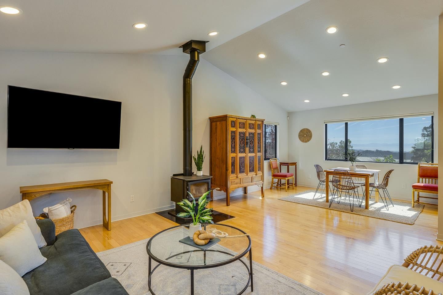 This bright and airy open-concept living and dining area features light hardwood flooring, a vaulted ceiling with recessed lighting, and a modern wood-burning stove as a central focal point. A large flat-screen television is mounted on the wall above a minimalist console table, while the dining space is positioned near expansive windows that offer a scenic view. The room is decorated with a blend of contemporary and rustic furniture, creating a warm and inviting atmosphere.