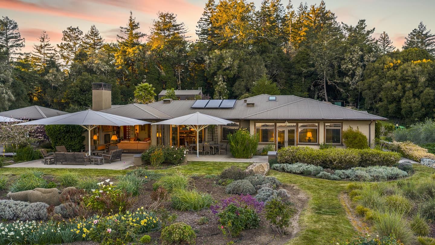 This stunning rear view of a single-story residence showcases a seamless blend of indoor-outdoor living with a spacious stone patio shaded by large, modern cantilever umbrellas. The home features a corrugated metal roof with integrated solar panels, large glass windows, and a warm, inviting interior visible from the outside. The property is set against a lush, forested backdrop and includes a beautifully landscaped garden with diverse plantings, creating a serene and picturesque atmosphere.