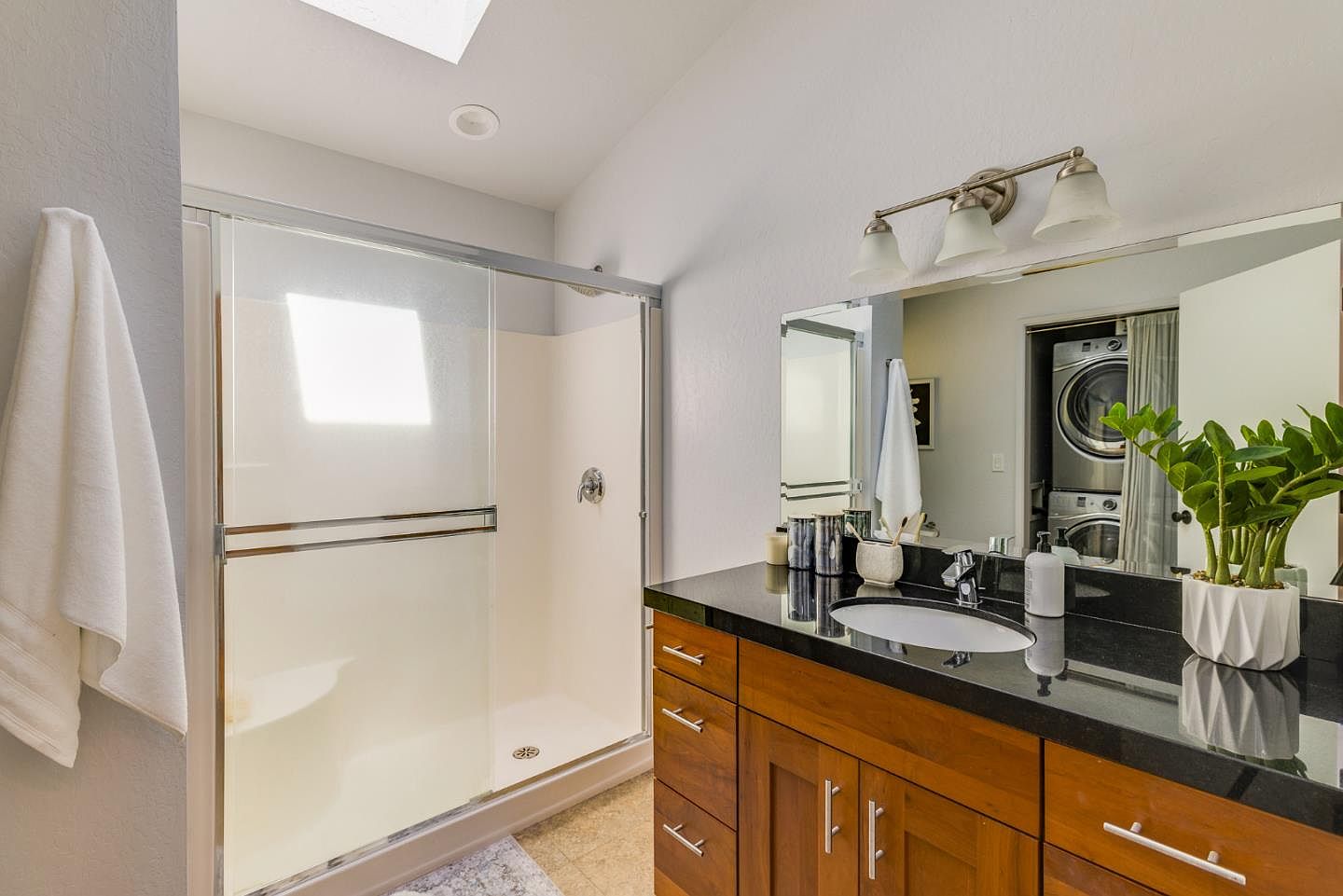 This bright and functional primary bathroom features a sleek vanity with a dark granite countertop, warm wood cabinetry, and a modern glass-enclosed shower. A large mirror reflects the space, including a glimpse of the laundry area, while a potted plant adds a touch of greenery to the clean, contemporary design. The room is well-lit by a combination of recessed lighting and a stylish three-bulb vanity fixture.