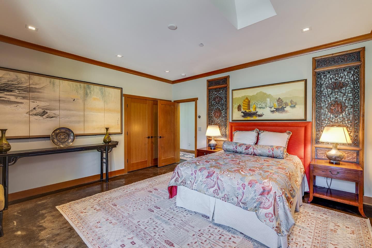 This elegant primary bedroom features a distinct Asian-inspired aesthetic, highlighted by intricate wooden wall panels and traditional landscape artwork. The room is anchored by a plush bed with a vibrant orange headboard, complemented by a patterned area rug and warm, ambient lighting from bedside lamps. The space exudes a serene and sophisticated atmosphere, blending cultural artistry with comfortable, high-end residential design.