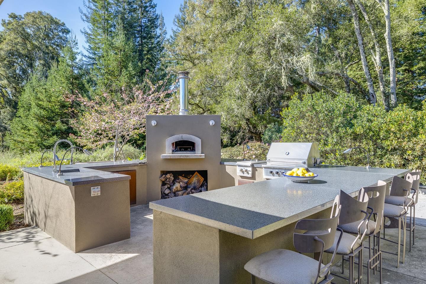 This outdoor kitchen area features a built-in pizza oven, a stainless steel grill, and a spacious countertop with bar seating, perfect for al fresco dining. The setup is nestled in a lush, wooded backyard setting, offering a serene and functional space for entertaining. The perspective is a wide, eye-level shot that captures the entire cooking and dining station against a backdrop of mature trees.