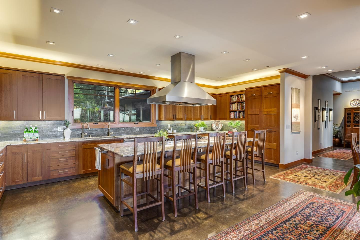 This spacious, warm-toned kitchen features rich wood cabinetry, a large granite-topped island with seating for five, and a prominent stainless steel range hood. The room is illuminated by recessed lighting and natural light from the large windows, creating an inviting atmosphere that blends traditional craftsmanship with modern functionality. The perspective is a wide-angle shot from the kitchen floor, showcasing the seamless flow between the cooking area and the adjacent hallway.