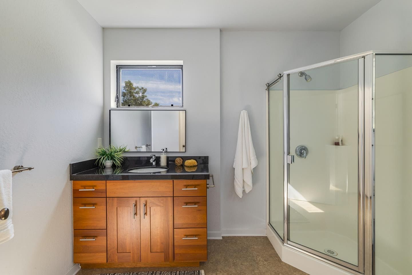 This primary bathroom features a warm wood vanity topped with a sleek black granite countertop and a modern undermount sink. To the right, a corner glass-enclosed shower with a white surround provides a clean, functional space, while a window above the vanity allows for natural light. The neutral wall color and simple design create a bright and inviting atmosphere.