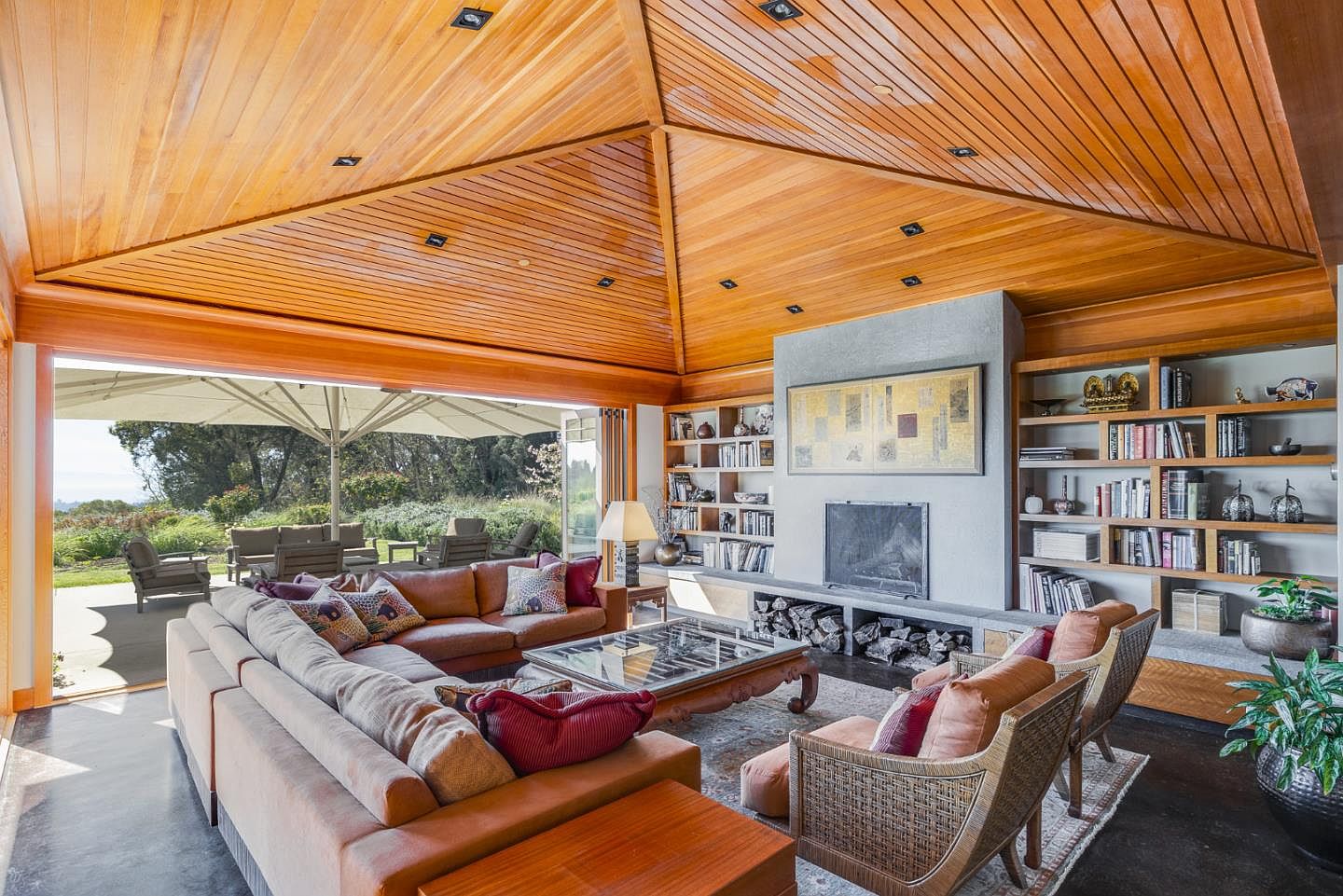 This spacious living room features a striking vaulted wood-paneled ceiling and a seamless indoor-outdoor transition through a massive sliding glass wall that opens to a patio. The room is anchored by a central fireplace with built-in shelving on either side, creating a warm, inviting atmosphere with a mix of rustic and contemporary design elements. The perspective is wide-angle, capturing the expansive seating area, the natural light flooding in from the garden, and the rich textures of the wood and stone finishes.