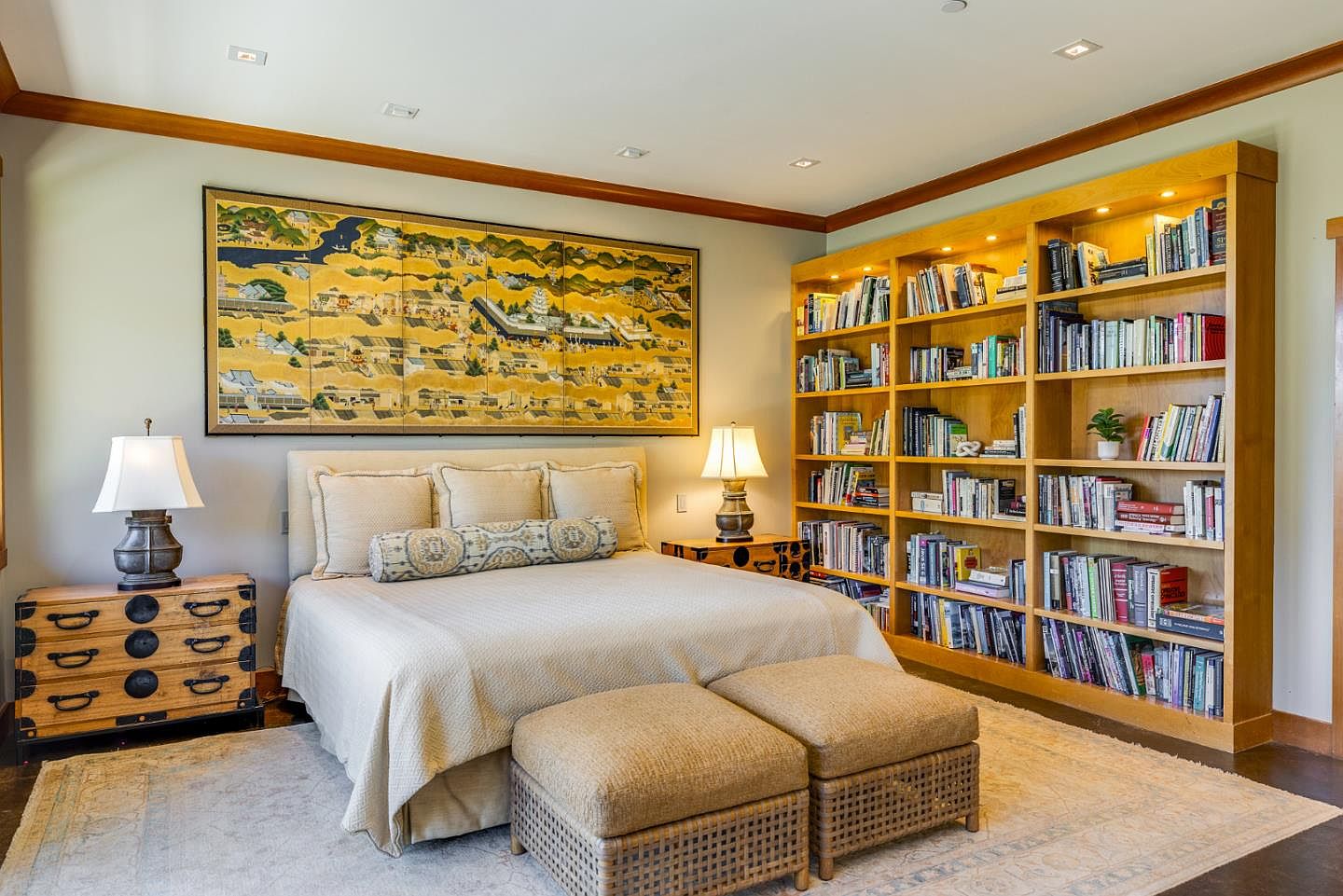 This serene primary bedroom features a neutral color palette, a plush bed with a decorative bolster pillow, and a large, ornate Japanese-style wall art piece as a focal point. A custom built-in wooden bookshelf provides ample storage and display space, while unique trunk-style nightstands add a touch of character. The room is grounded by a textured area rug and two matching ottomans at the foot of the bed, creating a sophisticated and cozy retreat.