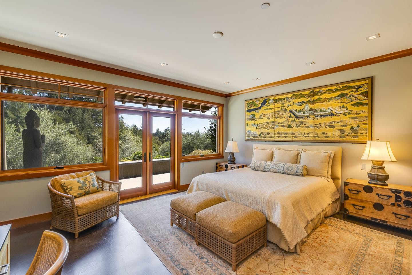 This serene primary bedroom features a neutral color palette, a large bed with a textured headboard, and a striking piece of Asian-inspired art above the bed. The room is bathed in natural light from the expansive windows and glass doors that lead to an outdoor deck, where a Buddha statue is visible. The space is furnished with a wicker armchair, matching ottomans, and unique wooden bedside tables, creating a tranquil and sophisticated retreat.