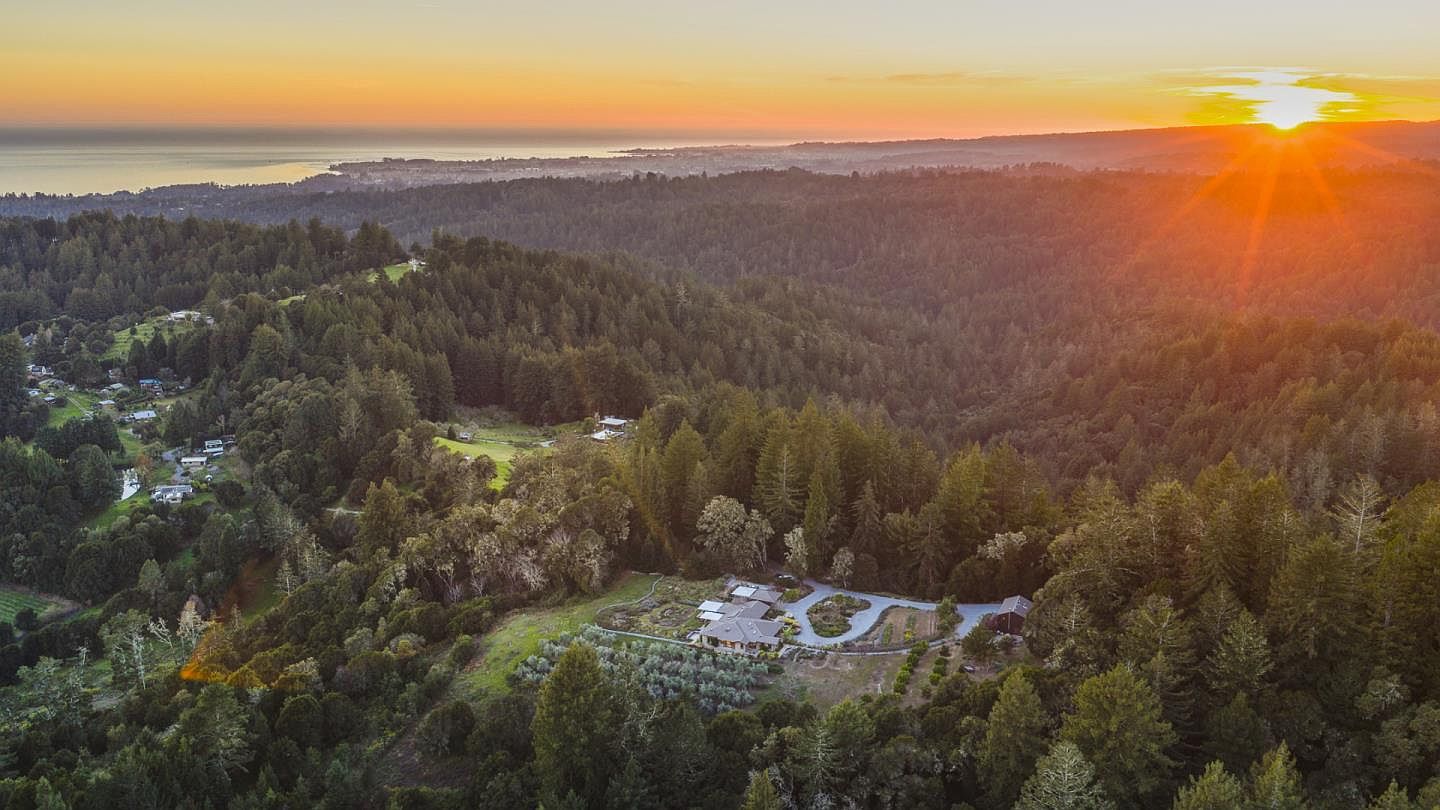 This stunning aerial view captures a secluded residential property nestled within a lush, dense forest, set against a breathtaking sunset over the distant coastline. The home features a well-maintained garden and orchard, highlighting a peaceful, private lifestyle surrounded by nature. The perspective emphasizes the expansive landscape and the serene, elevated position of the estate.
