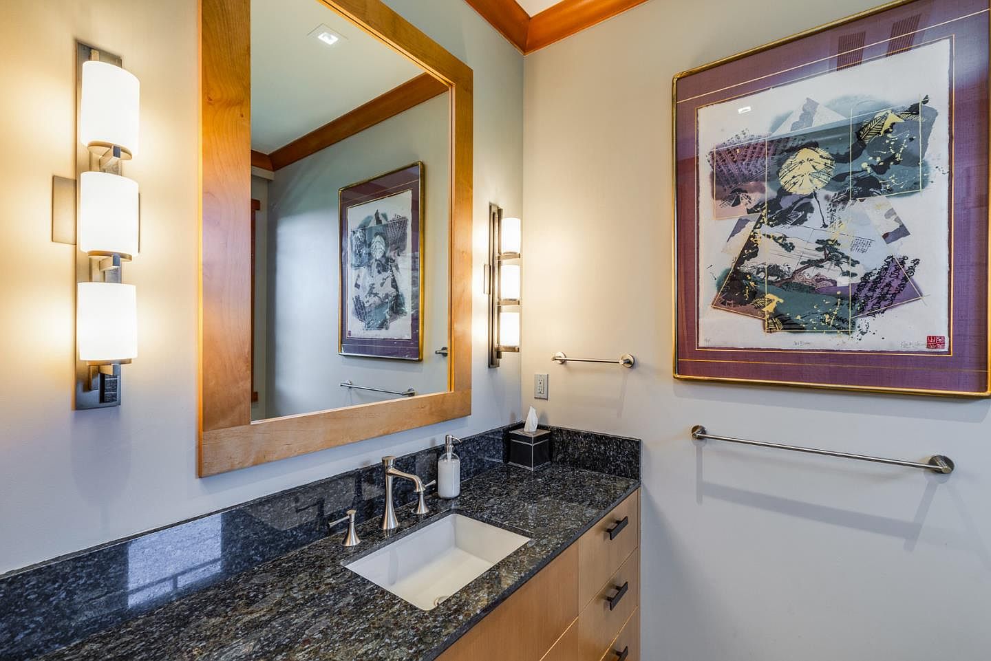 This elegant bathroom features a dark granite countertop with a clean white undermount sink and modern fixtures. A large mirror framed in warm wood is flanked by two stylish three-light sconces, while a framed piece of abstract art adds a sophisticated touch to the wall. The space is well-lit and organized, showcasing a contemporary design with high-quality finishes.