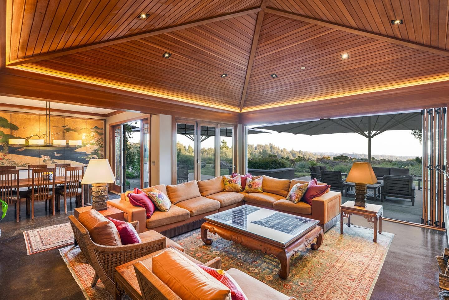 This spacious, open-concept living room features a dramatic vaulted wood-paneled ceiling with recessed lighting and a large, comfortable sectional sofa. The room seamlessly transitions to an outdoor patio through expansive folding glass doors, offering a panoramic view of the surrounding landscape. A traditional coffee table and patterned area rug anchor the space, creating a warm and inviting atmosphere that blends indoor comfort with natural beauty.