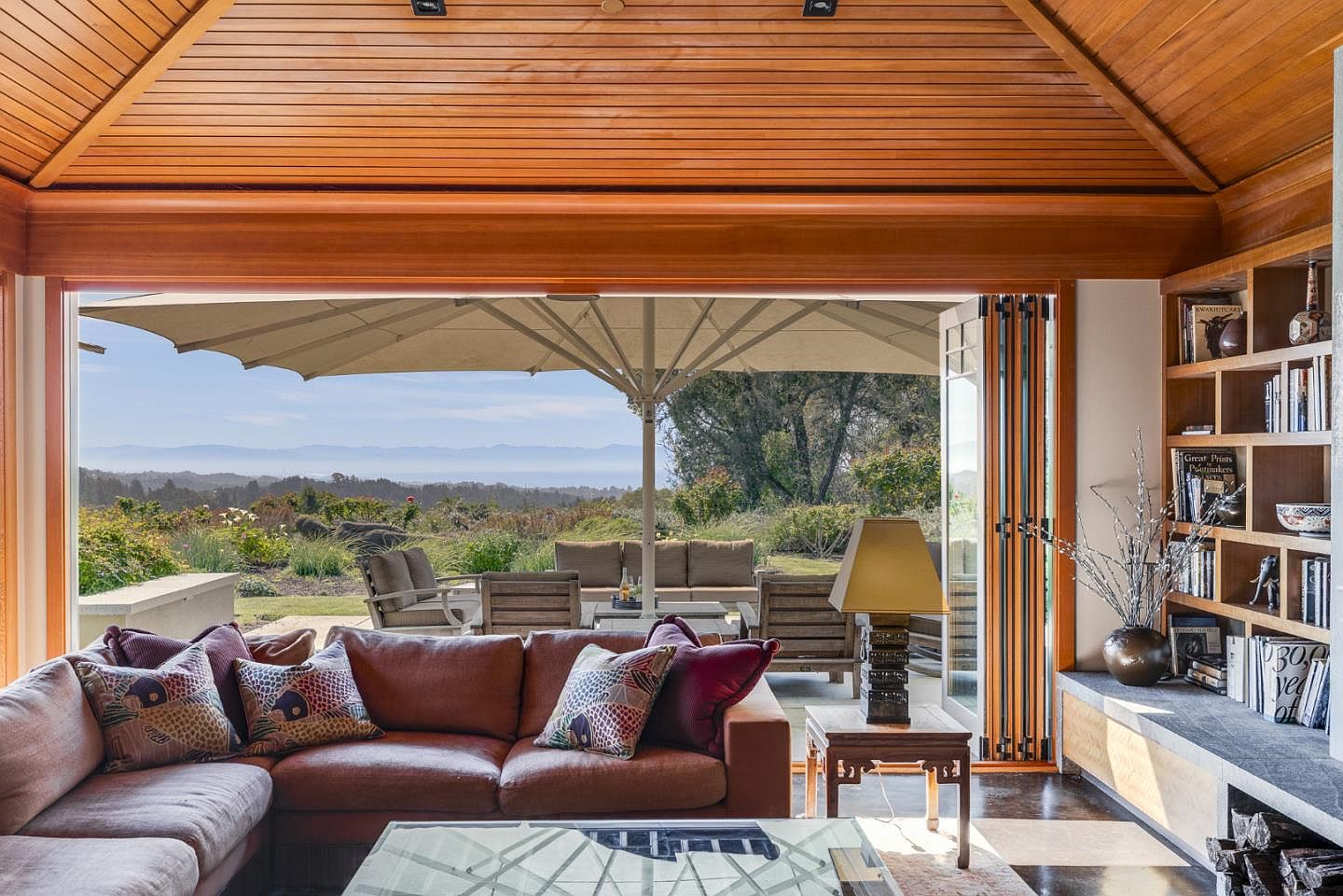 This inviting living room features a warm, wood-paneled vaulted ceiling and a comfortable sectional sofa adorned with patterned throw pillows. Large, folding glass doors open seamlessly to a spacious patio, blurring the lines between indoor and outdoor living while showcasing a breathtaking panoramic view of the rolling hills and distant mountains. The space is further enhanced by built-in shelving filled with books and decor, creating a cozy yet sophisticated atmosphere.