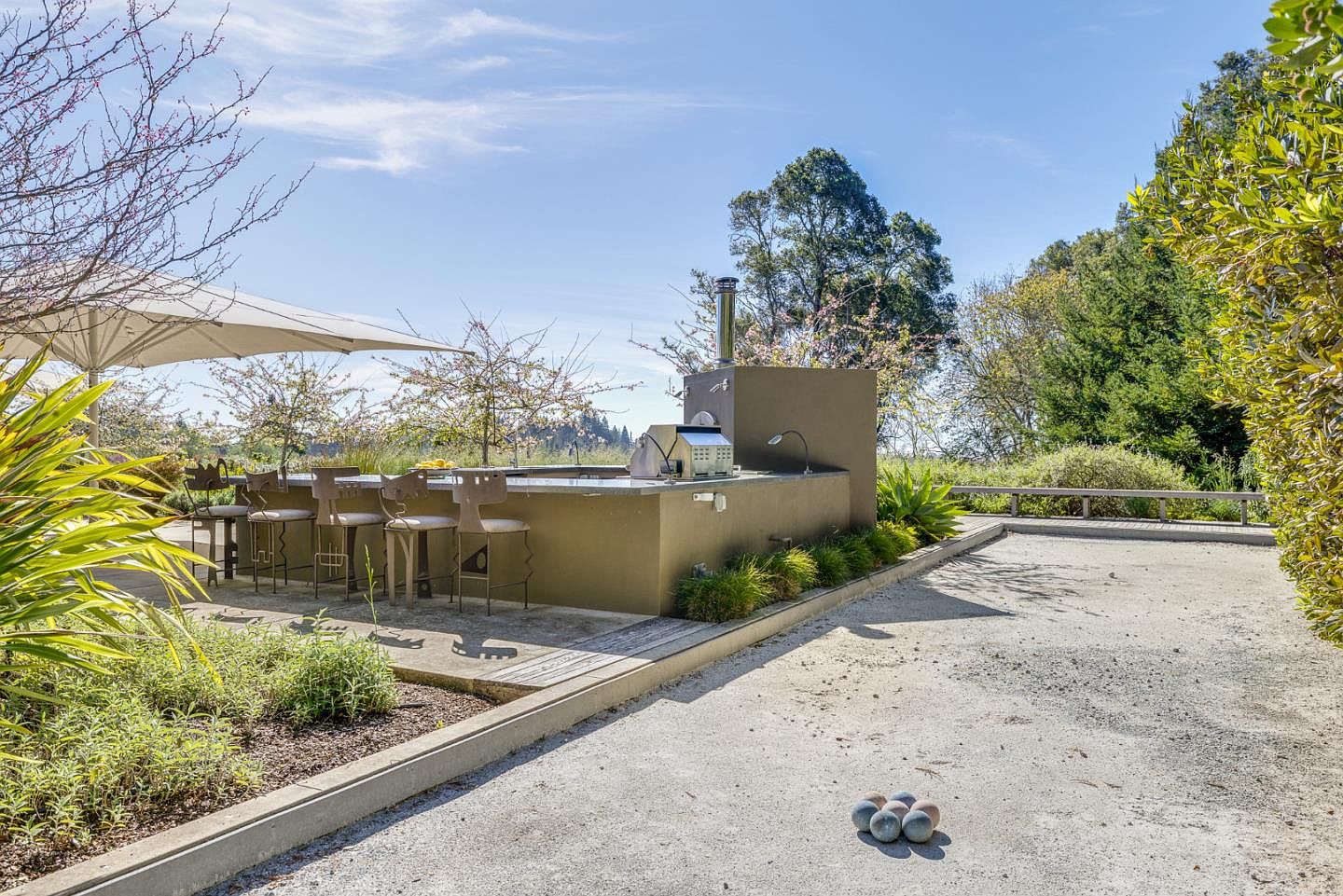 This outdoor living space features a sleek, modern concrete bar area equipped with a built-in grill and a chimney, perfect for alfresco entertaining. The patio is shaded by a large, contemporary umbrella and overlooks a gravel bocce ball court, creating a sophisticated yet relaxed atmosphere. The surrounding lush greenery and open sky provide a serene, resort-like backdrop for this high-end outdoor amenity.