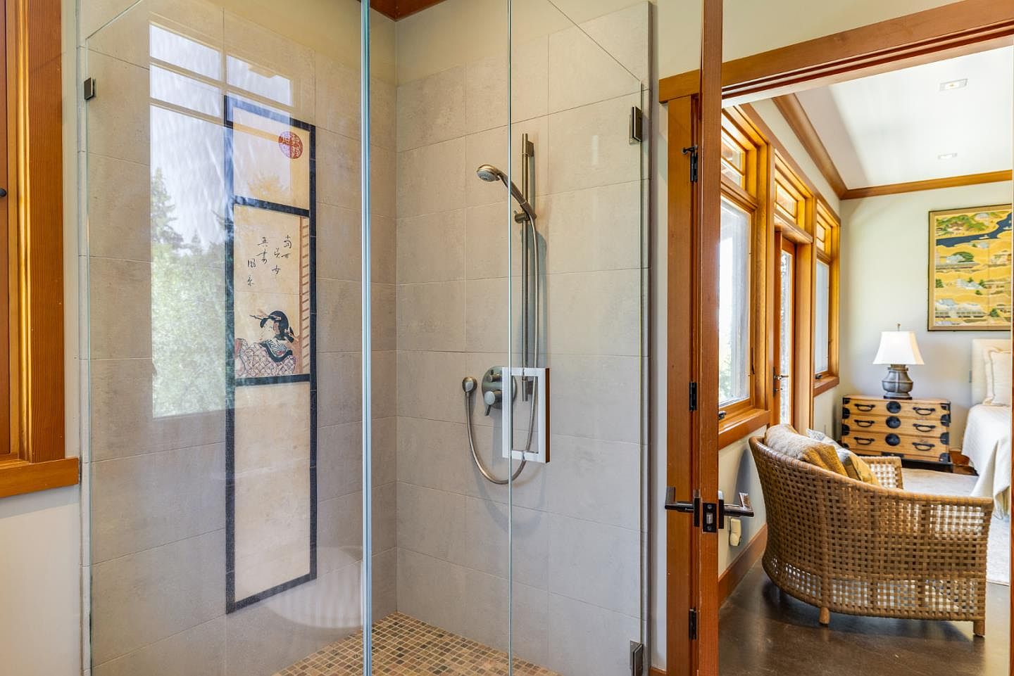 This image captures a sleek, modern walk-in shower with a glass enclosure, featuring neutral-toned wall tiles and a mosaic-tiled floor. Through the open doorway, a glimpse of a serene bedroom with a wicker chair and Asian-inspired artwork creates a cohesive, tranquil aesthetic. The perspective highlights the seamless transition between the functional bathroom space and the relaxing living area.