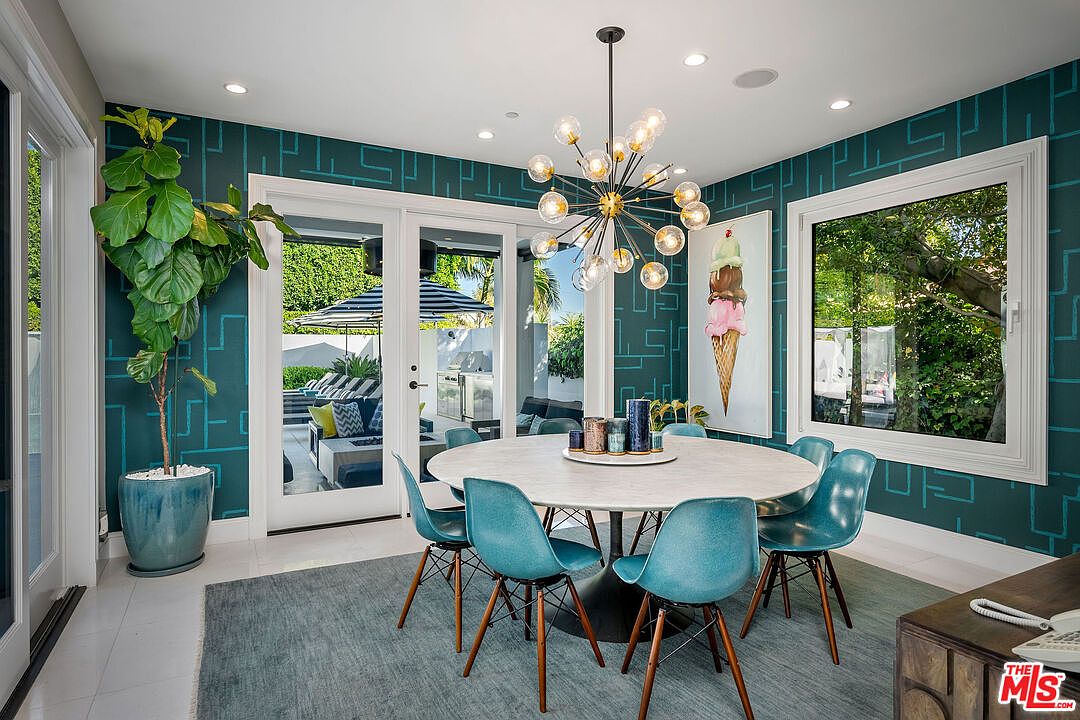 This vibrant dining room features a round table surrounded by mid-century modern style chairs in a bold teal hue, set against striking geometric patterned wallpaper. A large, multi-bulb sputnik-style chandelier serves as a focal point above the table, while a large piece of ice cream-themed wall art adds a playful touch. The room opens through glass doors to an outdoor patio area, creating a seamless transition between indoor and outdoor living spaces.