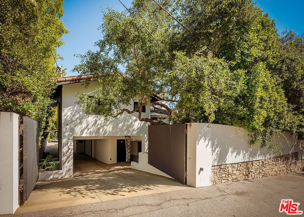 This exterior view captures the front of a modern, multi-level home nestled behind a private gate and lush, mature trees. The property features a clean, white-stucco facade with a recessed ground-level garage area and a stone-textured retaining wall along the street. The perspective is from the street level, emphasizing the home's secluded and gated entrance.