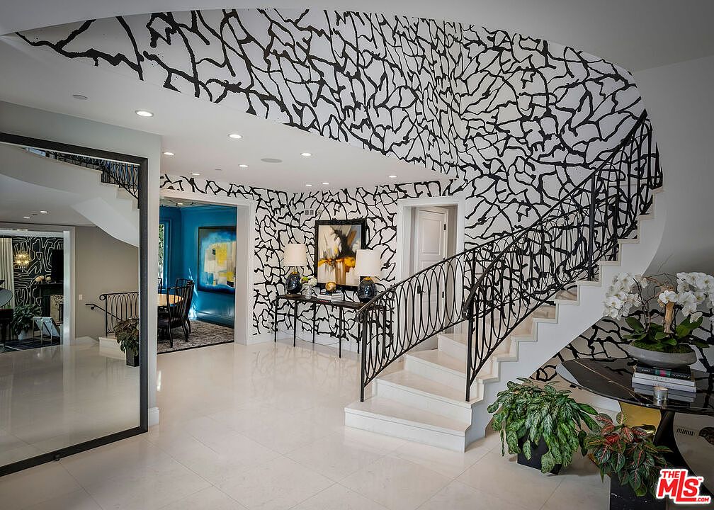 This grand foyer features a dramatic staircase with intricate black metal railings and a striking, bold black-and-white abstract wall pattern that extends across the ceiling. The space is anchored by polished light-colored stone flooring and includes a console table with lamps, a large mirror, and lush potted plants. A view into an adjacent room reveals a vibrant blue wall and contemporary artwork, creating a sophisticated and artistic atmosphere.