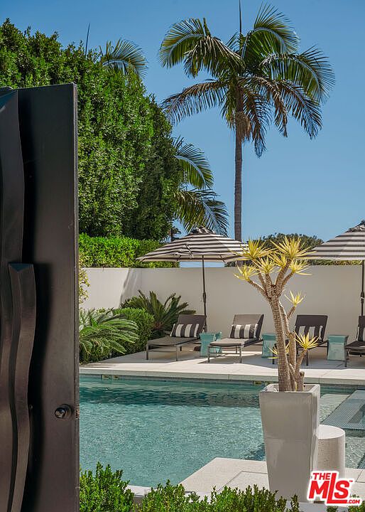 This view captures a serene swimming pool area framed by a dark, modern door frame in the foreground. The pool deck features several lounge chairs shaded by striped umbrellas, set against a clean white wall and lush green landscaping. A tall palm tree rises into the clear blue sky, creating a tranquil and luxurious outdoor retreat.