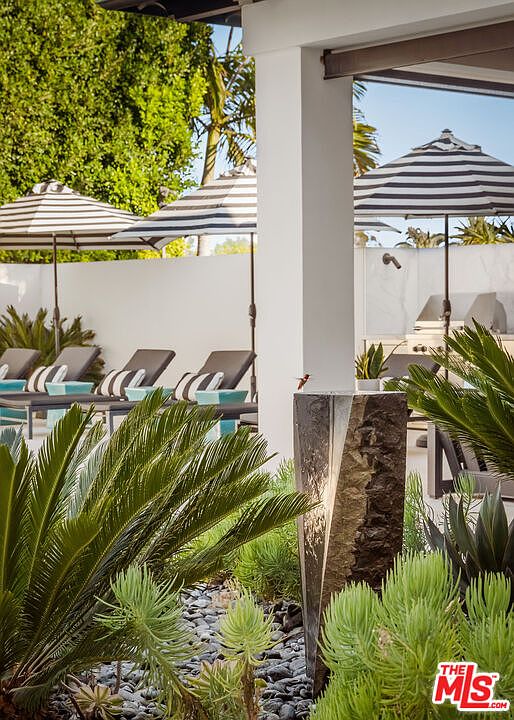 This outdoor patio space features a modern, minimalist aesthetic with a series of striped umbrellas providing shade over lounge chairs. A striking, angular stone water feature stands in the foreground, surrounded by lush, textured landscaping including cycads and succulents. The scene is captured from a low, cinematic perspective, emphasizing the clean lines of the architecture and the serene, resort-like atmosphere of the backyard.