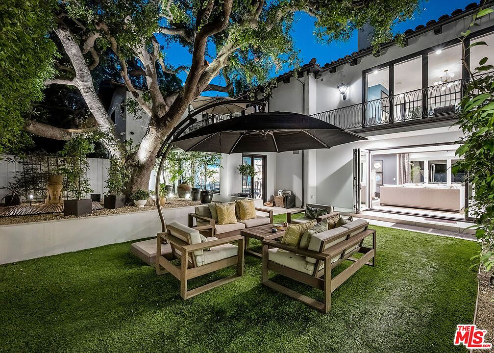 This evening shot captures a beautifully landscaped backyard featuring a synthetic lawn and a comfortable outdoor seating area shaded by a large cantilever umbrella. A mature, illuminated tree serves as a focal point, while the rear of the home showcases a modern aesthetic with large glass doors that open to the patio. The scene is framed from a low angle, emphasizing the seamless transition between the indoor living space and the private garden retreat.