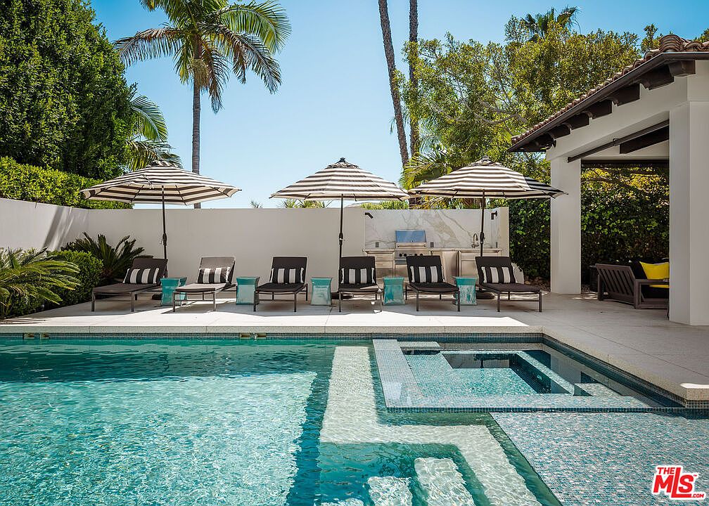 This luxurious outdoor living space features a pristine swimming pool with an integrated spa, set against a backdrop of lush palm trees and a clean white wall. A row of matching lounge chairs with striped umbrellas provides a comfortable seating area, complemented by a built-in outdoor kitchen station. The scene is captured from a low, eye-level perspective, emphasizing the seamless transition between the pool deck and the covered patio area.