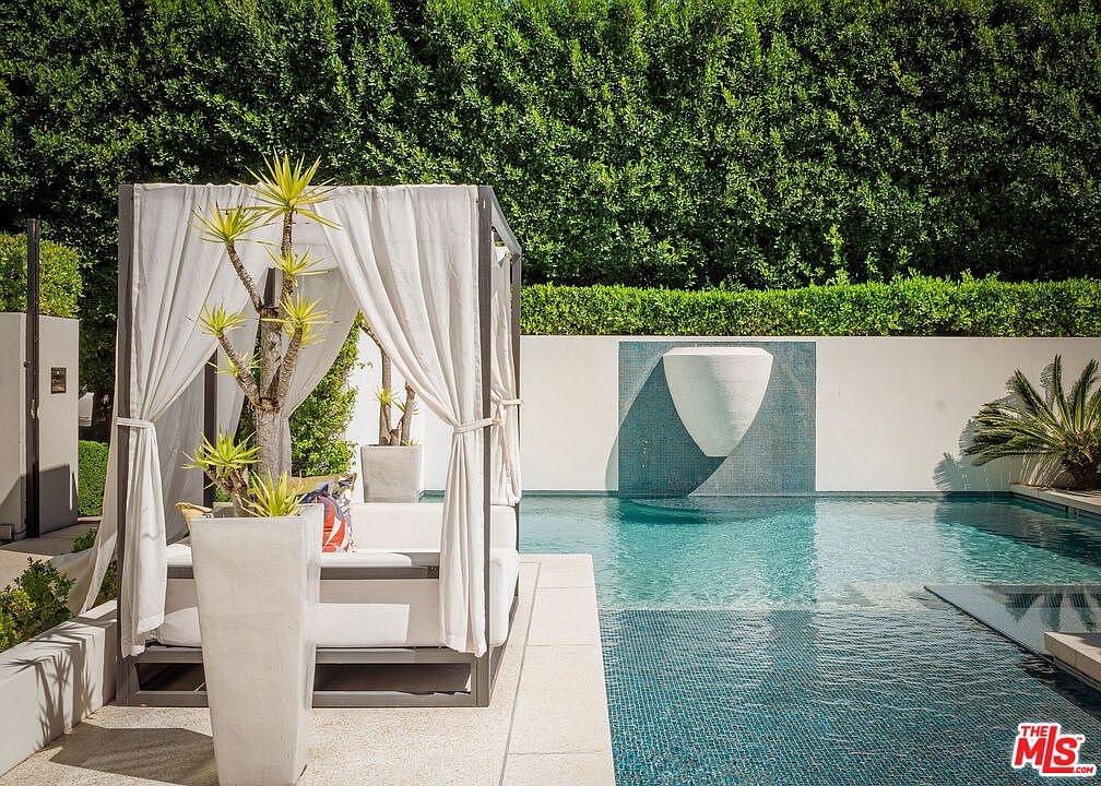 This luxurious outdoor space features a pristine swimming pool with a striking, modern water feature mounted on a white wall. A stylish cabana with white curtains and plush seating provides a shaded retreat, while a tall, sculptural plant in a large planter adds a touch of greenery to the poolside deck. The scene is framed by a dense, manicured hedge, creating a private and serene atmosphere perfect for relaxation.