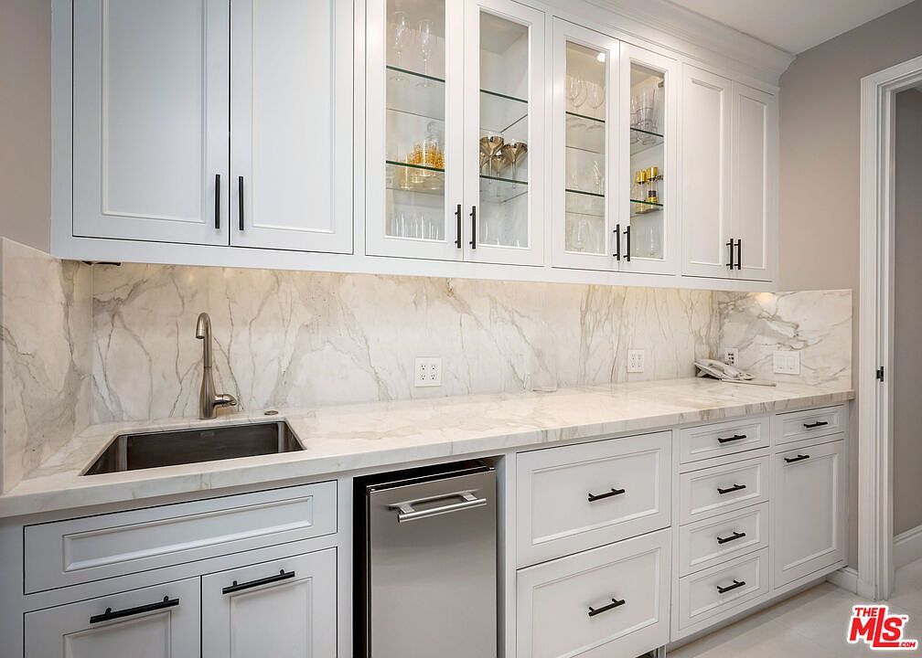 This elegant wet bar or butler's pantry features crisp white cabinetry with glass-front upper doors and sleek dark hardware. A light-toned stone countertop with dramatic veining extends across the workspace, complemented by a matching stone backsplash and an integrated stainless steel sink. The space is well-lit with under-cabinet lighting, creating a sophisticated and functional area for entertaining.