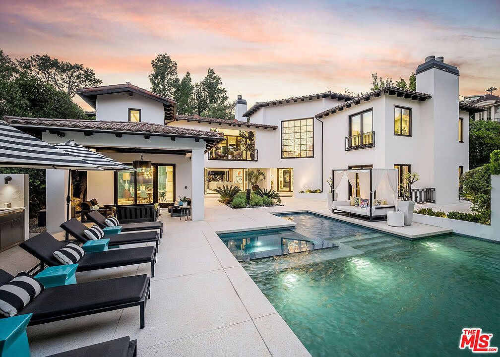 This luxurious rear exterior view showcases a sprawling white Mediterranean-style estate centered around a custom swimming pool and integrated spa. The expansive stone patio is furnished with multiple lounge chairs and a shaded cabana, creating an ideal setting for outdoor entertaining. The scene is captured at dusk, with the warm glow of interior lighting reflecting off the water and the soft colors of the sunset illuminating the architecture.