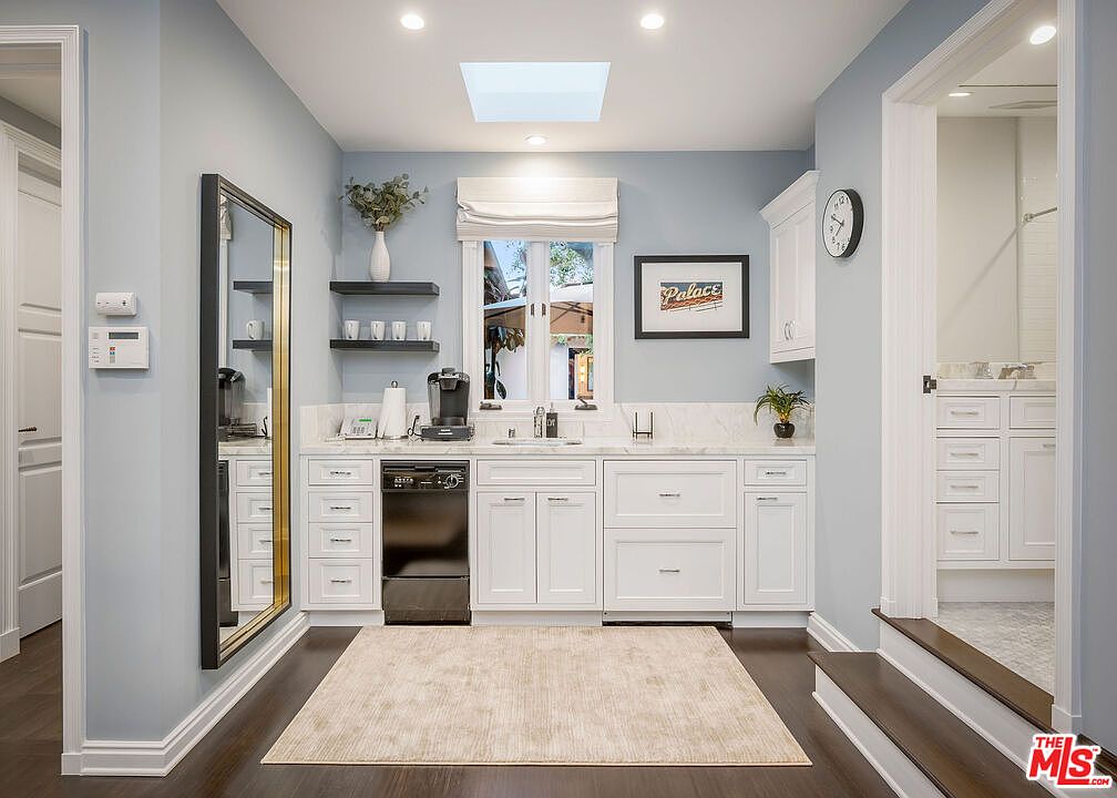 This bright and airy kitchenette features crisp white cabinetry, a dark dishwasher, and a light-toned stone countertop with an integrated sink. A large mirror with a black frame leans against the wall to the left, while a window above the counter provides natural light, complemented by a skylight overhead. The space is finished with dark wood flooring and a neutral area rug, creating a clean and functional transition area.