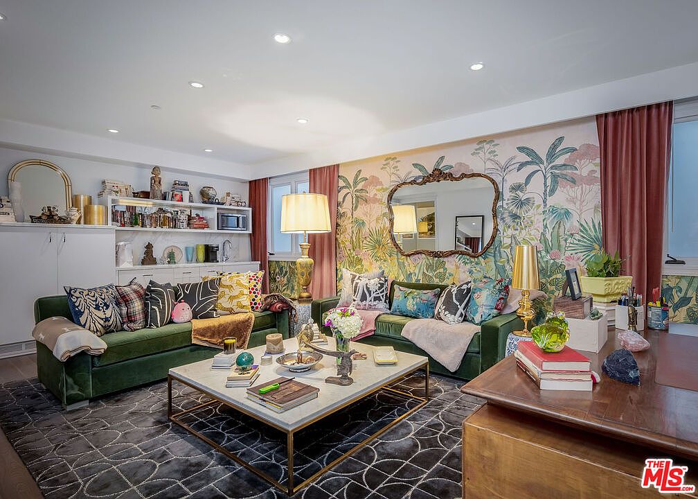 This vibrant living room features two plush green sofas arranged around a central stone-topped coffee table with a gold metal frame. The space is defined by a bold, botanical-themed wallpaper, a dark patterned area rug, and an ornate gold-framed mirror. The room is accessorized with numerous decorative pillows, lamps, and personal items, creating a maximalist and eclectic aesthetic.