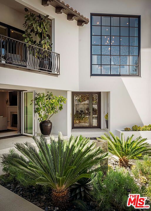 This elegant entryway features a crisp white stucco facade with a prominent grid-patterned window and a decorative iron-railed balcony above. The entrance is framed by a dark-toned, ornate metal door, while the foreground showcases a lush, manicured garden with cycad plants and dark river stones. The perspective is a ground-level shot looking toward the home, emphasizing the sophisticated Mediterranean-inspired architectural details and the seamless transition between indoor and outdoor living spaces.