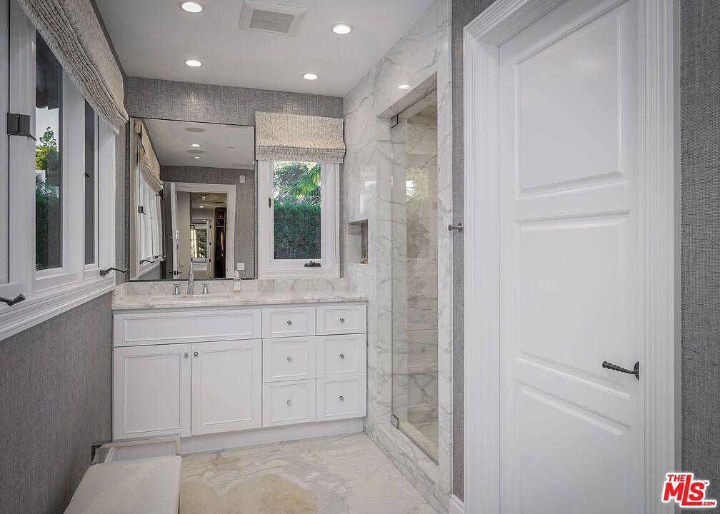 This bright and elegant bathroom features a white vanity with stone countertops, a large rectangular mirror, and a glass-enclosed shower with light-toned stone wall tiles. The room is accented by textured gray wallpaper and crisp white trim, creating a sophisticated and clean aesthetic. A window with a fabric roman shade allows natural light to fill the space, highlighting the polished stone flooring.