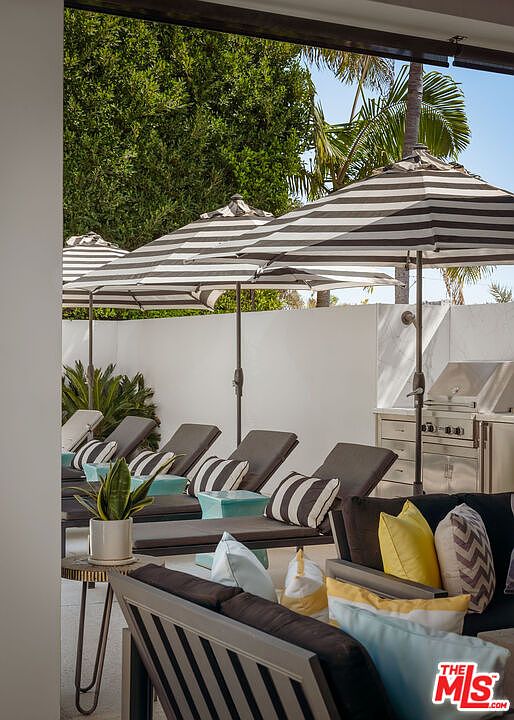 This outdoor patio space features a row of lounge chairs shaded by striped umbrellas, creating a comfortable and inviting relaxation area. A stainless steel outdoor grill is positioned against a clean white wall, while the foreground showcases a modern seating area with plush cushions in shades of yellow, blue, and grey. The perspective is from a covered area looking out, emphasizing a well-designed space perfect for outdoor entertaining.
